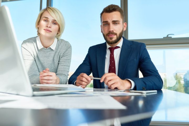 Portrait of handsome businessman and young woman looking at camera during meeting while discussing work using laptop at conference table in modern office against window